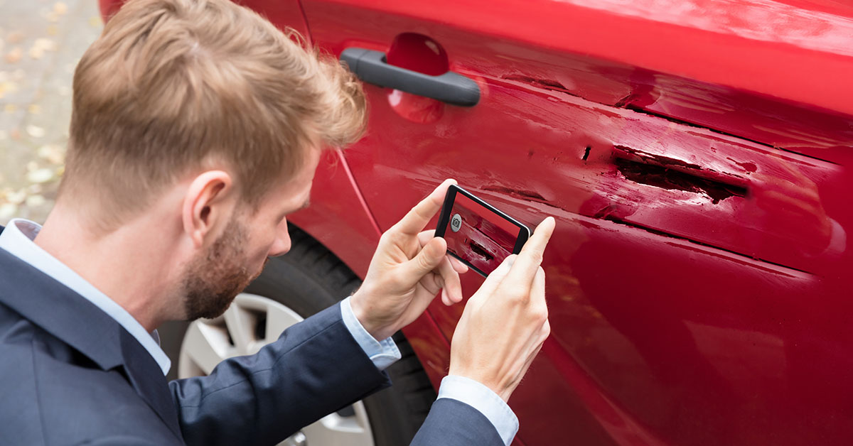 A man in a suit photographs his damaged car, capturing the aftermath of an unfortunate incident.