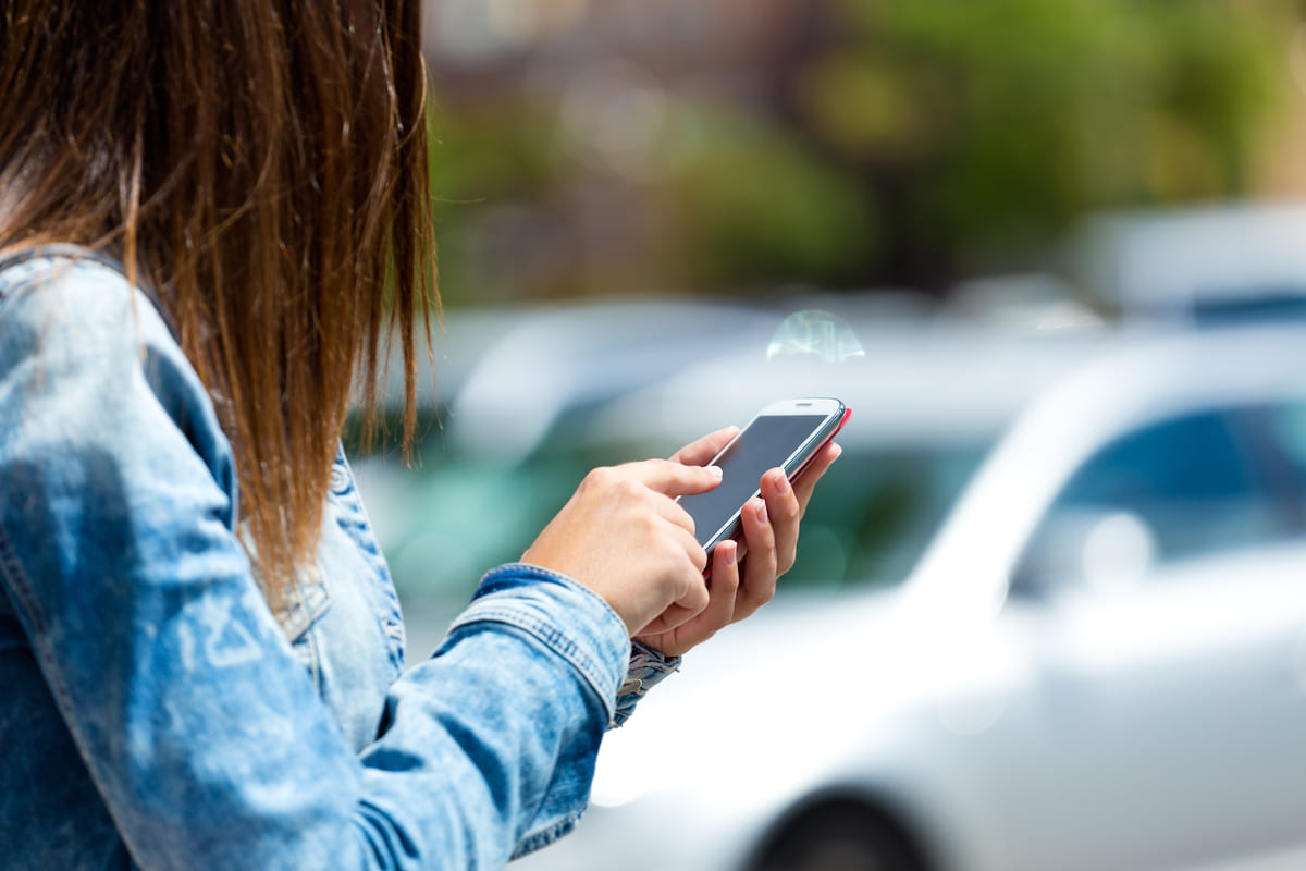 A woman stands in front of a car, using her cell phone with a focused expression.