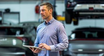 Fleet manager using a tablet in an automotive workshop with cars and a vehicle lift in the background