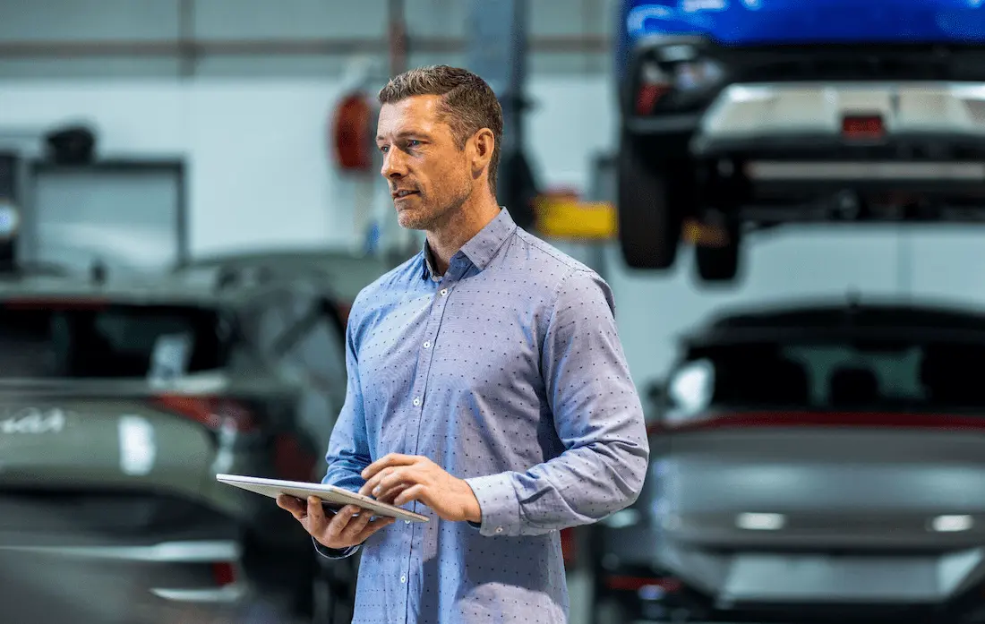 Fleet manager using a tablet in an automotive workshop with cars and a vehicle lift in the background