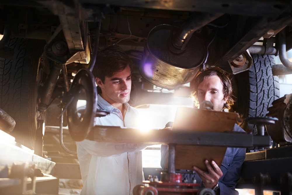 Automated driver communication tools for fleets: Two mechanics working underneath a raised vehicle while reviewing information on a clipboard in a workshop environment.