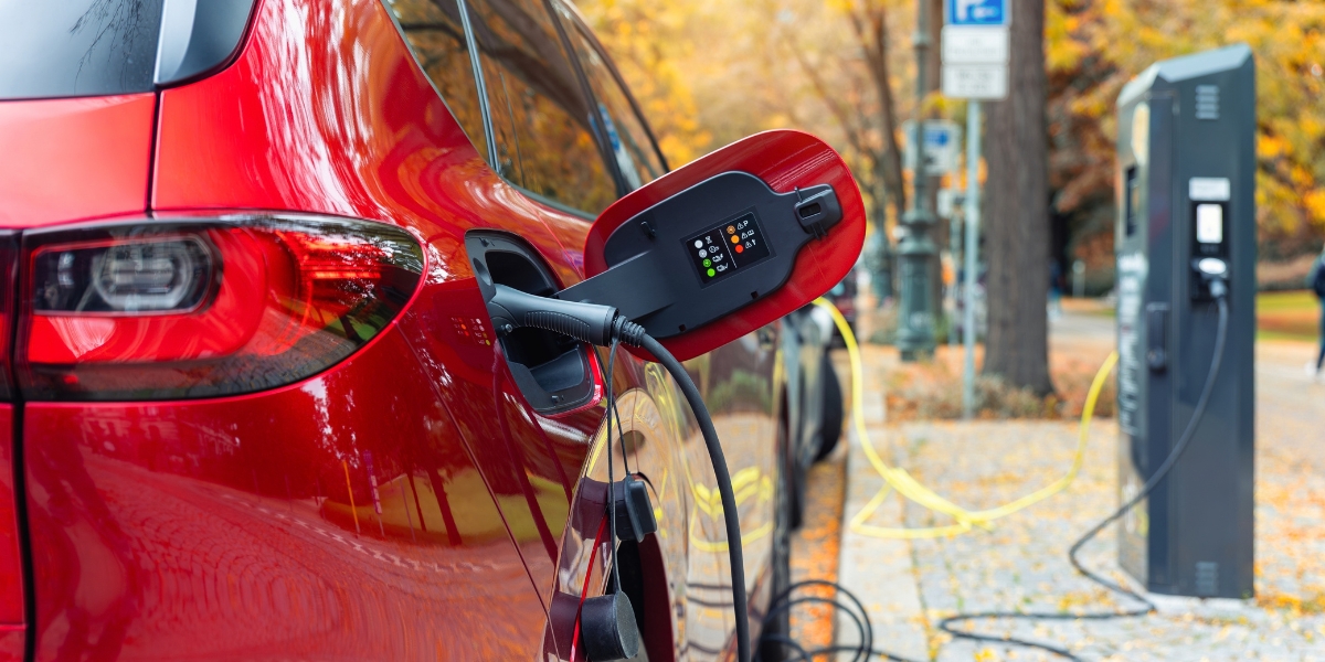 ev charging without a driveway: A red car plugged into a public charging point by a black cable, parked on a tree-lined street with autumn leaves on the pavement.