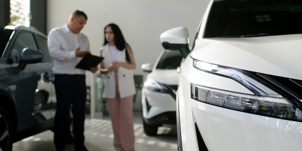 ev fleet transition plan: A manager with a clipboard talking to a woman in a car showroom, with a white SUV's headlight in focus in the foreground.