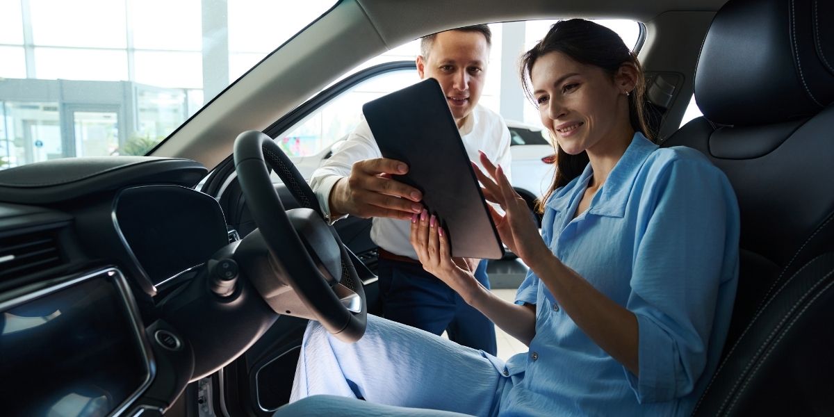 A smiling woman seated in a car reviewing a tablet with a salesman leaning in through the open door beside her.