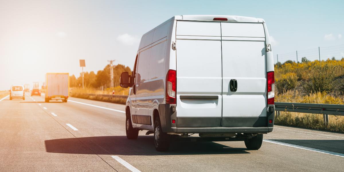 Fleet management van on road: A white panel van driving away from the camera along a sunlit motorway, with a lorry and other vehicles ahead in the left lane and fields alongside.