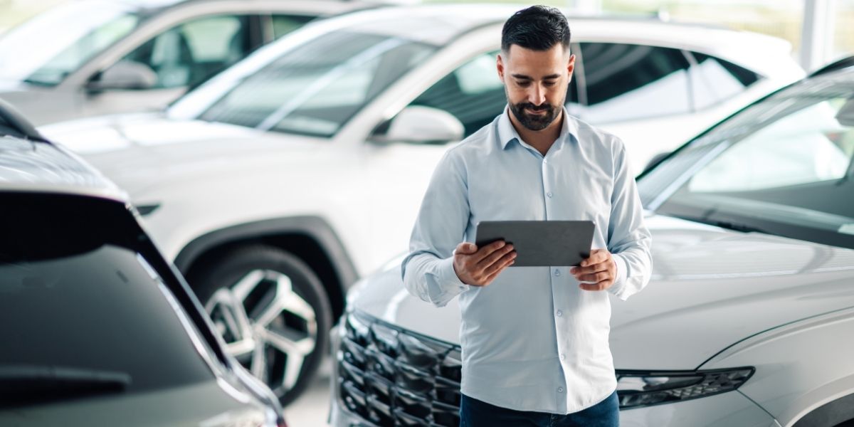 A man in a light blue shirt browsing a tablet whilst standing among a row of cars in a bright showroom.