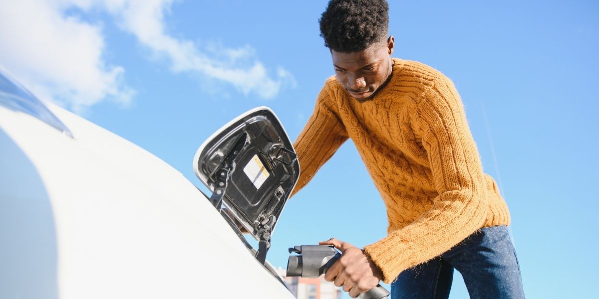 A man in a mustard yellow jumper plugging a charging cable into the charge port of a white electric car beneath a blue sky.