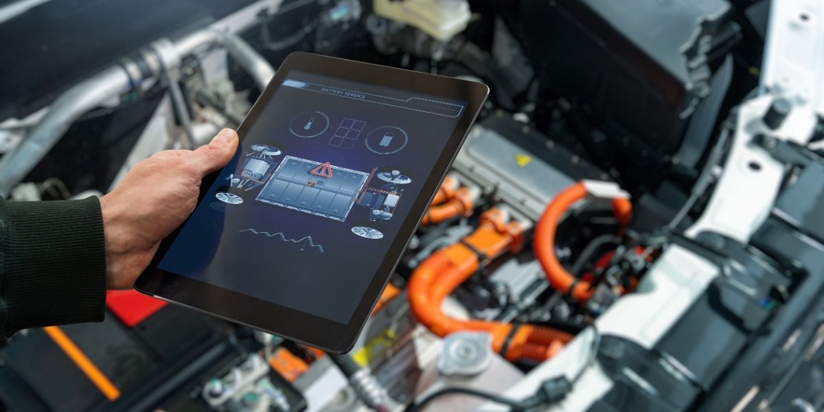 A hand holding a tablet displaying a battery service diagnostic interface, with a blurred electric vehicle engine bay showing orange high-voltage cables in the background.